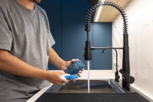 man washing glass with blue sponge black faucet sparkling dishes now