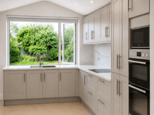 Linen Shaker kitchen in Malahide, Dublin with a large garden-facing window, marble-effect quartz worktops, integrated ovens and tall storage cabinets.