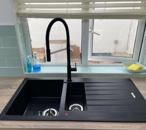 Black inset composite sink with integrated drainer and high-arc black tap beneath a window in a white shaker kitchen
