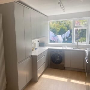 Sink positioned beneath a window in a light grey handleless U-shaped kitchen, with a built-in washing machine alongside