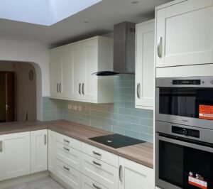 Cooking wall with a black glass hob and stacked double oven tower in a white shaker kitchen with a peninsula