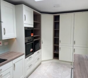 Full-height storage wall with a stacked double oven and open wood-grain shelving in a white shaker kitchen