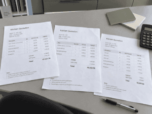 Three kitchen renovation quotes in euros laid out on a countertop beside cabinet samples, a calculator and a pen.