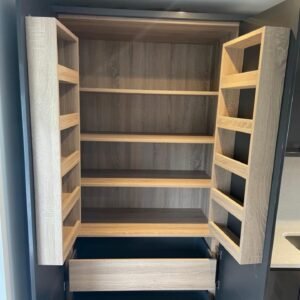 Open tall pantry cupboard with light oak-effect shelving, door racks and a base drawer in a gloss graphite kitchen