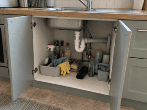 Open kitchen sink base cabinet showing plumbing pipes and cleaning supplies stored beneath the sink.